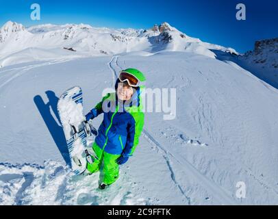 Wide mountain panorama and cute little smiling boy hold snowboard in hand aside, view from above with mask off Stock Photo