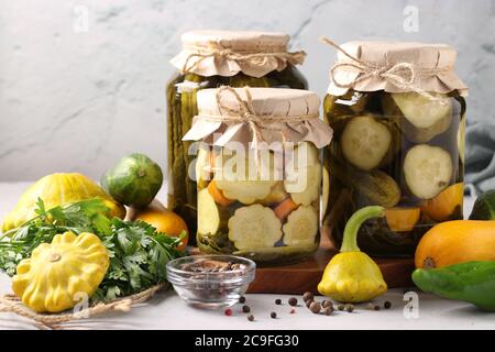 Homemade pickled patissons, cucumbers and zucchini in glass jars and fresh ingredients on a light gray background in still life, Closeup Stock Photo