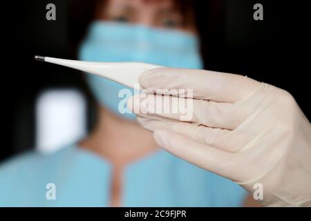 Coronavirus symptoms, woman in medical mask measures body temperature. Doctor looks at digital thermometer in her hand, concept of cold and flu Stock Photo