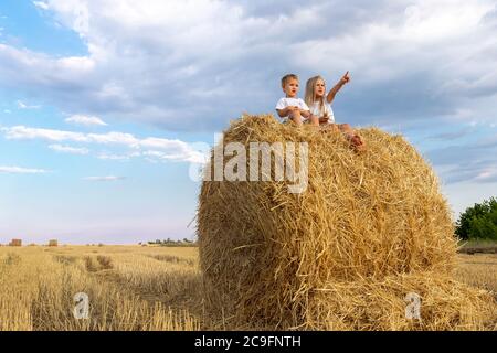 Two cute adorable caucasian siblings enjoy having fun sitting on top over golden hay bale on wheat harvested field near farm. Happy childhood and Stock Photo