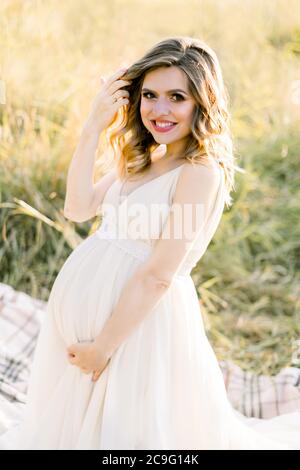 Pretty caucasian pregnant woman in white dress sitting on a picnic blanket on a meadow at sunset. Summer evening in nature. Happy maternity and Stock Photo