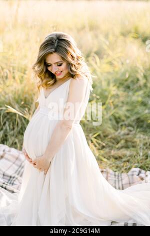Pretty caucasian pregnant woman in white dress sitting on a picnic blanket on a meadow at sunset. Summer evening in nature. Happy maternity and Stock Photo