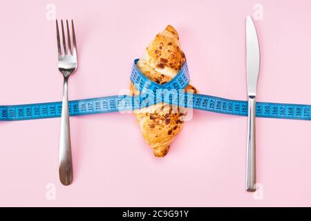 A baked almond croissant wrapped in a measuring tape on a pink background. The concept of diet, anti-obesity, weight loss by counting calories and avo Stock Photo