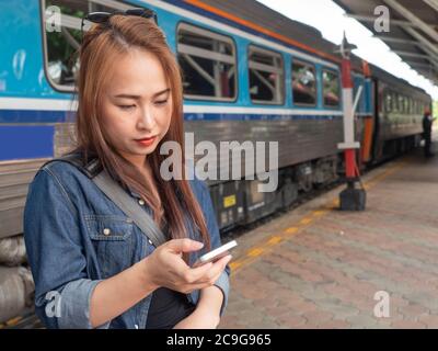 Asian women using smartphones at the train platform with the train in the background. Young Asian lady searching for information on a smartphone at th Stock Photo