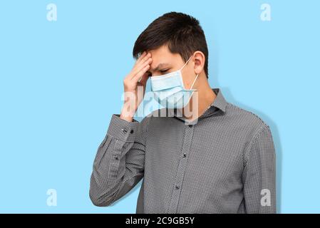 Portrait of teenage boy in protective medical mask with symptoms of covid-19 on light blue background. Teenager holding his head suffering from headac Stock Photo