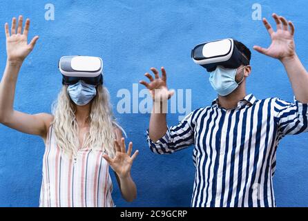 Young couple wearing face surgical mask having fun with virtual reality glasses during outbreak - Millennial people using innovated vr Stock Photo