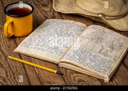 Vintage expedition journal on a rustic picnic table with a cup of tea and straw hat - handwriting and drawing in travel log from 1974 paddling trip in Stock Photo