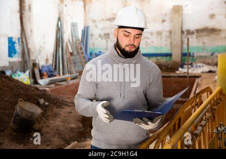 Focused young engineer planning renovation works at building site, looking at papers Stock Photo