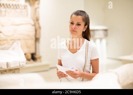 Young female local tourist reading guidebook with exhibition program looking at sculptures in historical museum Stock Photo