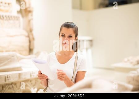 Young female local tourist reading guidebook with exhibition program looking at sculptures in historical museum Stock Photo