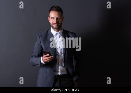 A smiling handsome young businessman in a suit using his smartphone in front of a dark grey background in a studio. Stock Photo
