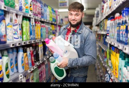 Cheerful bearded male standing with household chemicals in hands among shelves in supermarket, satisfied with purchases Stock Photo