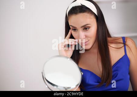 beauty, skin care and people concept - smiling young woman in hairband touching her face and looking to mirror at home bathroom Stock Photo