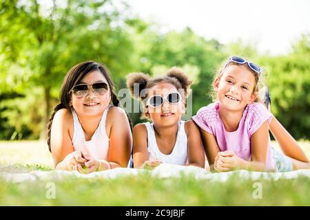 Girls wearing sunglasses and lying side by side on blanket in park, smiling, portrait. Stock Photo