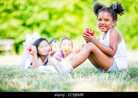 Children lying in park, girl sitting and holding apple in foreground. Stock Photo