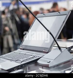 Detail from a business presentation. Microphone and laptop on the speech podium, blurred crowd in the background. Stock Photo
