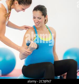 Pregnant woman exercising, sitting on fitness ball, instructor helping. Stock Photo