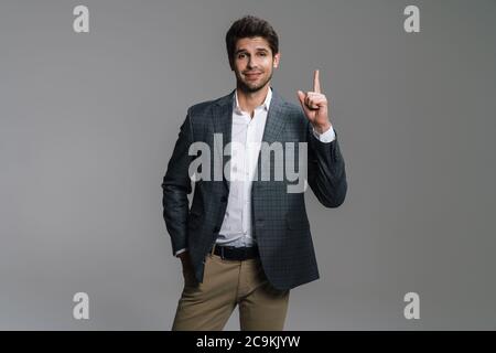 Photo of cheerful businessman in jacket smiling and pointing finger upward isolated over grey wall Stock Photo