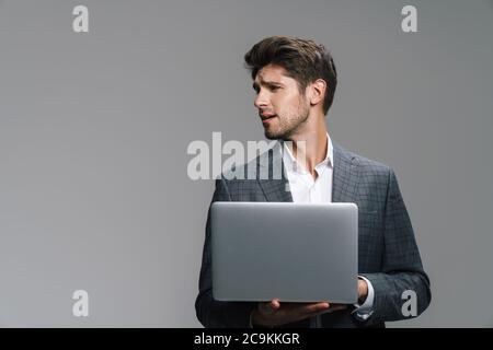 Photo of confused young businessman working with laptop isolated over grey wall Stock Photo