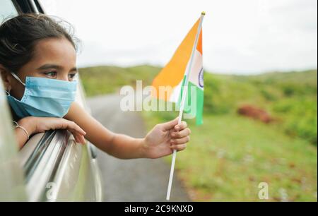 Young girl kid with medical mask holding Indian flag in moving car window - Concept of celebrating Independence or republic day during coronavirus or Stock Photo