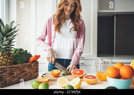 Charming caucasian woman with red hair and freckles is slicing fruits with a knife and making juice at home Stock Photo