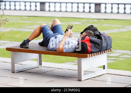 Stockholm, Sweden - July 31, 2020: A young man is lying on a park bench with a bag as a pillow in the city hall park and using his mobile phone. Stock Photo
