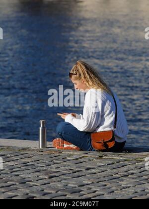 Stockholm, Sweden - Juluy 31, 2020: A blonde young woman wearing a white knitted sweater and blue jeans using a mobile phone is sitting in a tailoring Stock Photo