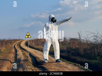 Man in white coverall, gas mask and gloves standing on village road in the field next to radiation sign pointing and looking away, on sunny day under blue sky. Concept of ecology, biological hazard. Stock Photo