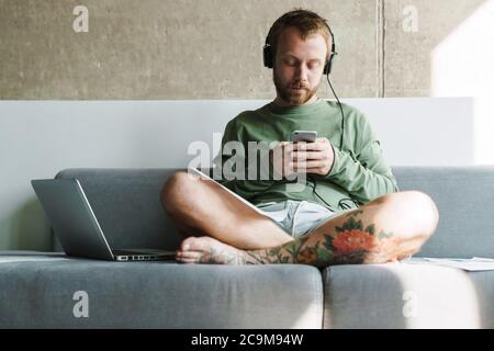Photo of young focused man using mobile phone and headphones while working with laptop on sofa at home Stock Photo