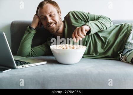 Photo of young cheerful man with beard watching movie on laptop while eating popcorn at home Stock Photo