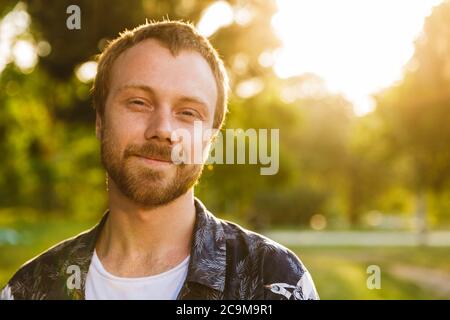 Image of young caucasian bearded man smiling and looking at you while walking in summer green park Stock Photo