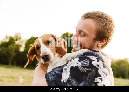 Image of young caucasian bearded man smiling and sitting with his beagle dog in summer park Stock Photo