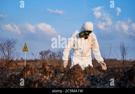 Environmentalist in gas mask and protective suit staring at burnt field in despair. Male ecologist looking at damaged grass and soil after fire. Yellow biohazard sign and blue sky on background Stock Photo