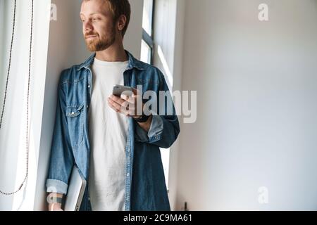 Photo of pleased bearded man using mobile phone and holding laptop while leaning on wall indoors Stock Photo
