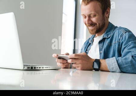 Photo of smiling handsome man playing online game on cellphone while working with laptop at table indoors Stock Photo