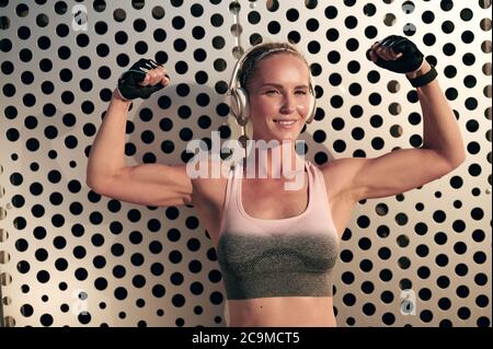 Smiling athletic woman in headphones and sportswear is showing muscles in her arms and listens to music, strong girl with perfect body