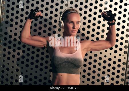 Athletic woman in headphones and sportswear is showing muscles in her arms and listens to music