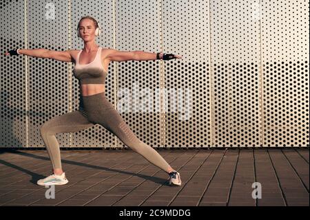 Yoga virabhadrasana II warrior pose. Woman in headphones practicing yoga against wall, urban background Stock Photo