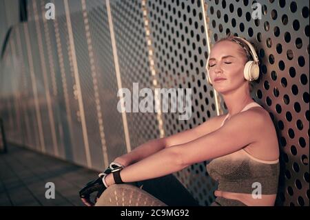 Athlete female runner in sportswear relaxing sitting closing eyes and listening to music outdoor in front of wall Stock Photo