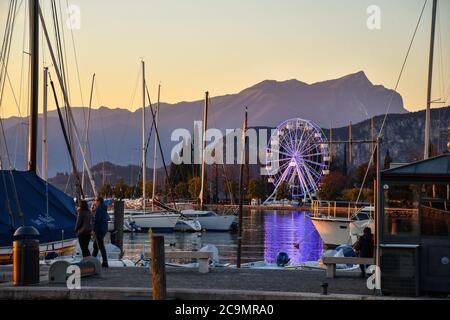 View of the harbor on the shore of Lake Garda with the illuminated big wheel and the mountains in the background at sunset, Bardolino, Veneto, Italy Stock Photo