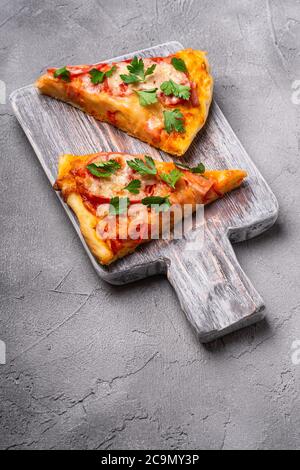 Hot pizza slices with mozzarella cheese, ham, tomato and parsley on wooden cutting board, stone concrete background, angle view Stock Photo