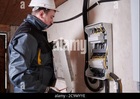 Electrical engineer in work uniform checking electric box with wires and power fuse switches. Man technician wearing white safety helmet. Concept of electricity and safe electrical power supply. Stock Photo