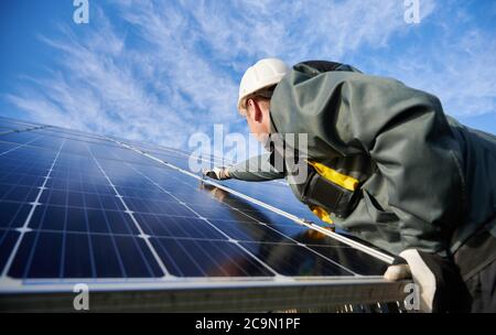 Side view worker wearing uniform, working gloves and helmet under blue sky on background. Technician setting a shiny new solar battery with help of hex key. Concept of alternative green energy Stock Photo