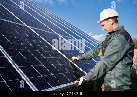 Side view of man technician in safety helmet repairing photovoltaic solar module. Professional electrician worker in gloves maintaining solar photovoltaic panel system. Concept of alternative energy Stock Photo