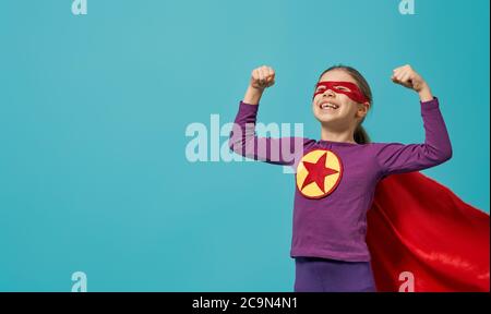 Little child is playing superhero. Kid on the background of bright blue wall. Girl power concept. Stock Photo