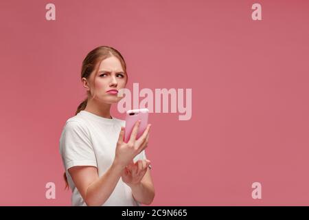 Confused young redhead woman holding pink smartphone.Embarassed girl has problem with broken not working mobile phone.Receiving bad news or email,spam Stock Photo