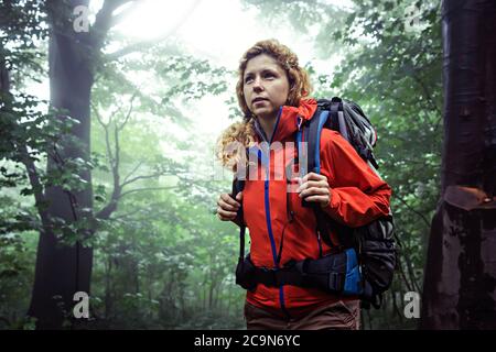 Portrait of a female trekker, wearing outdoor clothing and hiking backpack, trekking through deep, misty forest. Stock Photo