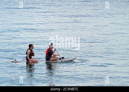 Two women sitting & paddle boarding on paddle board in calm tranquil water on River Thames estuary close to shoreline Southend on Sea Essex England UK Stock Photo