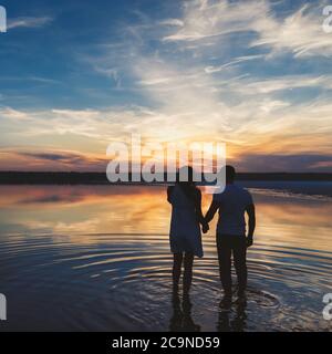 Portrait, silhouette of happy couple watching the colourful bright sunset standing in large lake, reflection in the water, holding hands Stock Photo