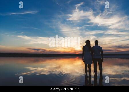 Portrait, silhouette of happy couple watching the colourful bright sunset standing in large lake, reflection in the water, holding hands Stock Photo